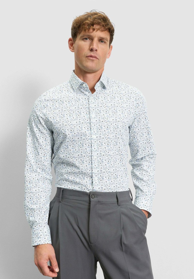 Man wearing a white long-sleeve shirt with a small floral pattern and gray high-waisted trousers, standing with one hand in pocket.
