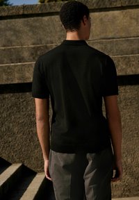 Schwarzes Kurzarm-Poloshirt mit glatter Textur, kombiniert mit grauen Streifenhosen; Ansicht von hinten vor einem steinernen Hintergrund.
