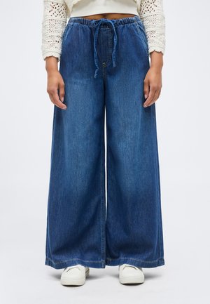 Pantalon large en denim bleu avec taille élastique et cordon de serrage, porté avec des baskets blanches et un haut en crochet crème à manches longues.
