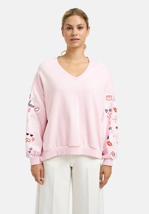 Woman wearing light pink V-neck sweatshirt with colorful embroidered symbols on sleeves and white pants, standing against white background.
