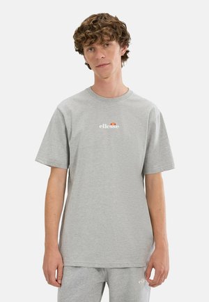 Junger Mann mit lockigem Haar trägt ein hellgraues ellesse T-Shirt und passende Shorts, steht vor einem schlichten weißen Hintergrund.