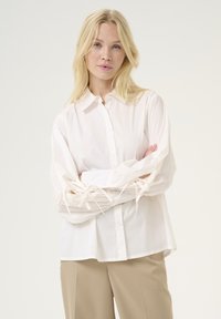 Camicetta bianca con maniche lunghe caratterizzata da dettagli annodati sui polsini, realizzata in un tessuto leggero e morbido. Design semplice.