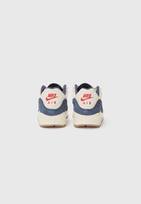 Nike sportbačiai su tamsiai mėlyna zomšo viršutine dalimi ir grietinėlės spalvos akcentais. Su raudona ženklinimo etikete ant kulno ir tekstūruotu tinkleliu aplink apykaklę.