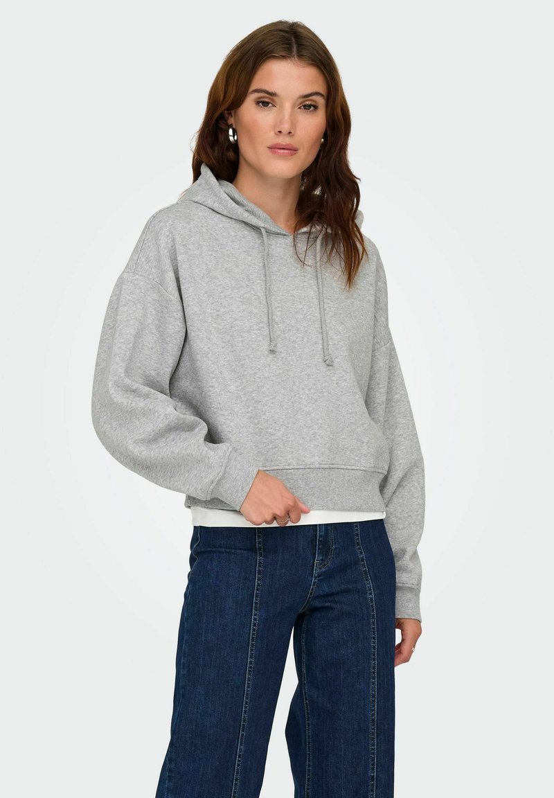 Femme portant un sweat à capuche gris clair et un jean bleu foncé, se tenant contre un fond blanc uni, avec une main près de sa taille.