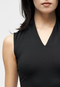 Γυναίκα με αμάνικο μαύρο τοπ με V λαιμόκοψη, εμφανίζεται από τους ώμους έως τα χείλη, με φόντο ένα απλό λευκό φόντο.