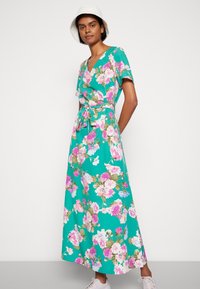 Vestido maxi em tecido teal com um padrão floral, apresentando flores rosas e brancas. Design em V com mangas curtas e cintura amarrada.