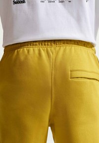 Gros plan sur un pantalon taille élastique jaune moutarde avec une poche passepoilée unique à l'arrière, porté avec un t-shirt blanc à motif rentré à l'intérieur.