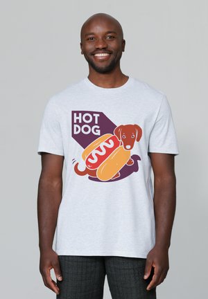 Lächelnder Mann trägt ein weißes T-Shirt mit einem Cartoon-Dackel, der als Hot Dog verkleidet ist, und dem Text "HOT DOG" auf hellem grauem Hintergrund.