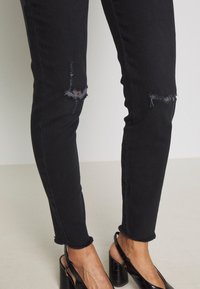 Jean skinny noir avec des déchirures aux genoux usés, des ourlets effilochés et une texture lisse. Associé à des chaussures noires à talons.