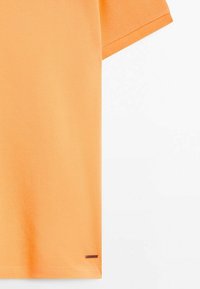 Polo en coton orange à manches courtes avec une petite étiquette métallique rectangulaire près de l'ourlet inférieur sur fond blanc.