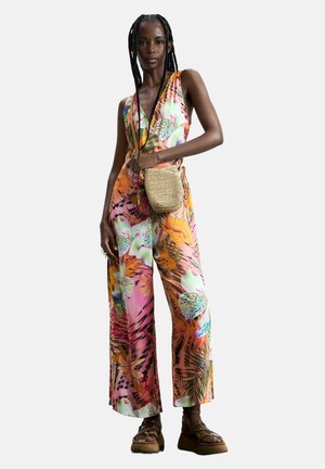 Kvinde med lange fletninger iført en ærmeløs jumpsuit med blomsterprint, der holder en vævet crossbody-taske og har brune plateau-sandaler på.