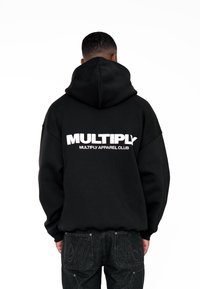 Felpa nera oversize con un ampio logo bianco "MULTIPLY" sul retro, dotata di cappuccio con coulisse e tessuto morbido. Indossata con jeans di denim scuro.