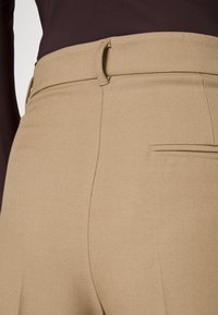 Nærbillede af beige skræddersyede bukser med bæltesløjfer og baglommer, båret med en mørkebrun top.