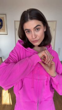 Mladá žena s tmavými vlasy ve světlém růžovém mikině uvnitř, upravující si rukáv s neutrálním výrazem.