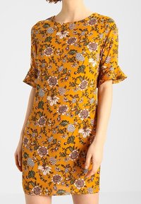 Žena v hořčicově žlutých šatech ke kolenům s krátkými volánovými rukávy a květinovým vzorem v bílé, hnědé, zelené a fialové barvě.