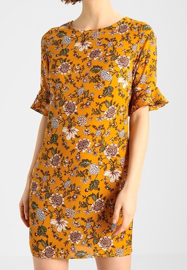 Žena v hořčicově žlutých šatech ke kolenům s krátkými volánovými rukávy a květinovým vzorem v bílé, hnědé, zelené a fialové barvě.