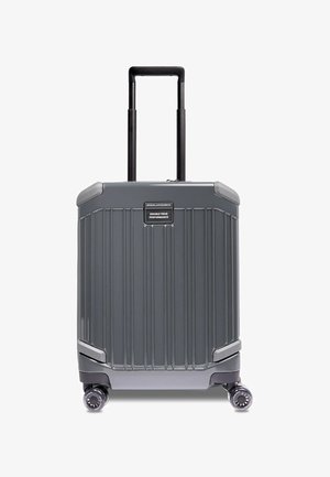 Valise rigide grise avec rainures verticales, poignée télescopique double, quatre roues et une étiquette de marque sur le devant.