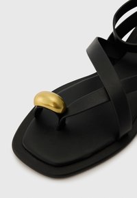 Černé kožené sandály se čtveratou špičkou a zkříženými pásky. Vyznačují se zlatým metalickým akcentem na špičce. Hladká textura a minimalistický design.