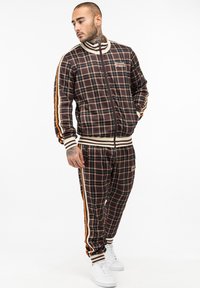 Brown plaid tracksuit with white and orange stripes, featuring a zip-up jacket and matching pants, made of smooth fabric with ribbed cuffs.