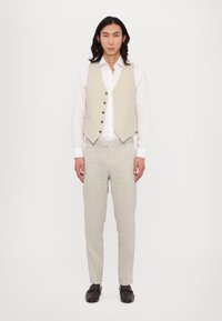 Beige Weste mit Knöpfen über einem weißen Hemd, kombiniert mit hellgrauen Hosen. Das Outfit zeichnet sich durch ein maßgeschneidertes Design und eine glatte Textur aus.