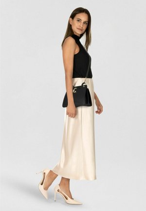 Top nero a coste senza maniche, gonna midi in raso color crema, tacchi a spillo beige con dettagli a taglio, borsa nera con dettagli dorati.