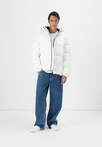 Hvid pufferjakke med høj krave, lynlåslukning og kontrastfarvet mørk hætteforing. Kombinationen med løse blå jeans og hvide sko.