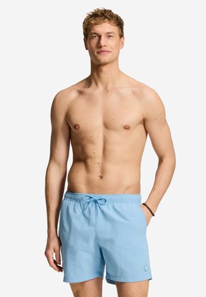 Mees lokkis blondide juustega, seljas helesinised ujumispüksid, seisab valge tausta ees ühe käe taskus.