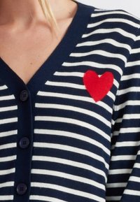 Cardigan a righe blu e bianche con scollatura a V, dotato di un cuore ricamato rosso sul lato sinistro e bottoni scuri lungo la parte anteriore.