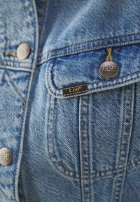 Žep modre denim jakne z metalnimi gumbi in tekstilno etiketom, na kateri je napisana blagovna znamka "Lee".