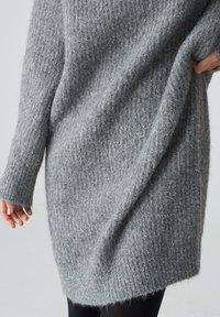 Grauer strickpullover mit langen Ärmeln, weicher Textur, lockerer Passform und einem leicht gewölbten Saum. Der Stoff weist ein sichtbares, fusselartiges Muster auf.