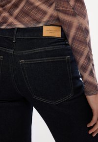 Temno modri kavbojki z gladko teksturo, ki imajo na pasu rjavo usnjeno nalepko z napisom "popolni kavbojki". Klasična zasnova z petimi žepi.