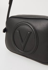 Svart läder crossbody-väska med en texturerad yta, med en framträdande präglad rund logotyp och en platt design. Justerbar rem ingår.