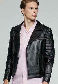 Zwarte leren motorjack met diagonale rits, schouderepauletten en geribbelde accenten op de mouwen. Draagt over een lichtroze shirt.