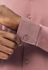 Asmuo užsegantis mygtuką ant mauviško ilgos rankovės marškinio, parodant artimą plano vaizdą su rankomis ir audinio tekstūra.