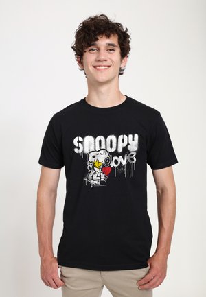 Jeune homme souriant aux cheveux bouclés portant un t-shirt noir avec Snoopy qui embrasse Woodstock avec les mots "Snoopy love" et un cœur rouge.