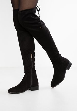 Bottes noires en daim au-dessus du genou avec fermeture éclair latérale, petit talon, bord clouté à l'avant, portées avec des collants noirs sur fond blanc.