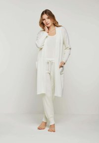 Weißer langer Cardigan mit offenem Front, gerippten Ärmeln und zwei seitlichen Taschen, kombiniert mit einem weißen, figurbetonten Oberteil und passenden Hosen.