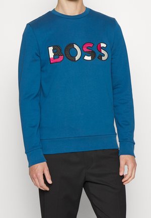 Mann trägt einen blauen Sweatshirt mit langen Ärmeln und dem Wort „BOSS“ in mehrfarbigen, strukturierten Buchstaben auf der Brust sowie eine schwarze Hose.
