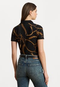 Polo nero con motivo a catena dorata, maniche corte e colletto, abbinato a jeans blu sbiaditi a vita alta.
