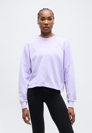 Γυναίκα με πλεξούδες φοράει ένα φαρδύ φούτερ ανοιχτό μωβ και μαύρο κολάν, στέκεται μπροστά σε ένα απλό γκρι φόντο.