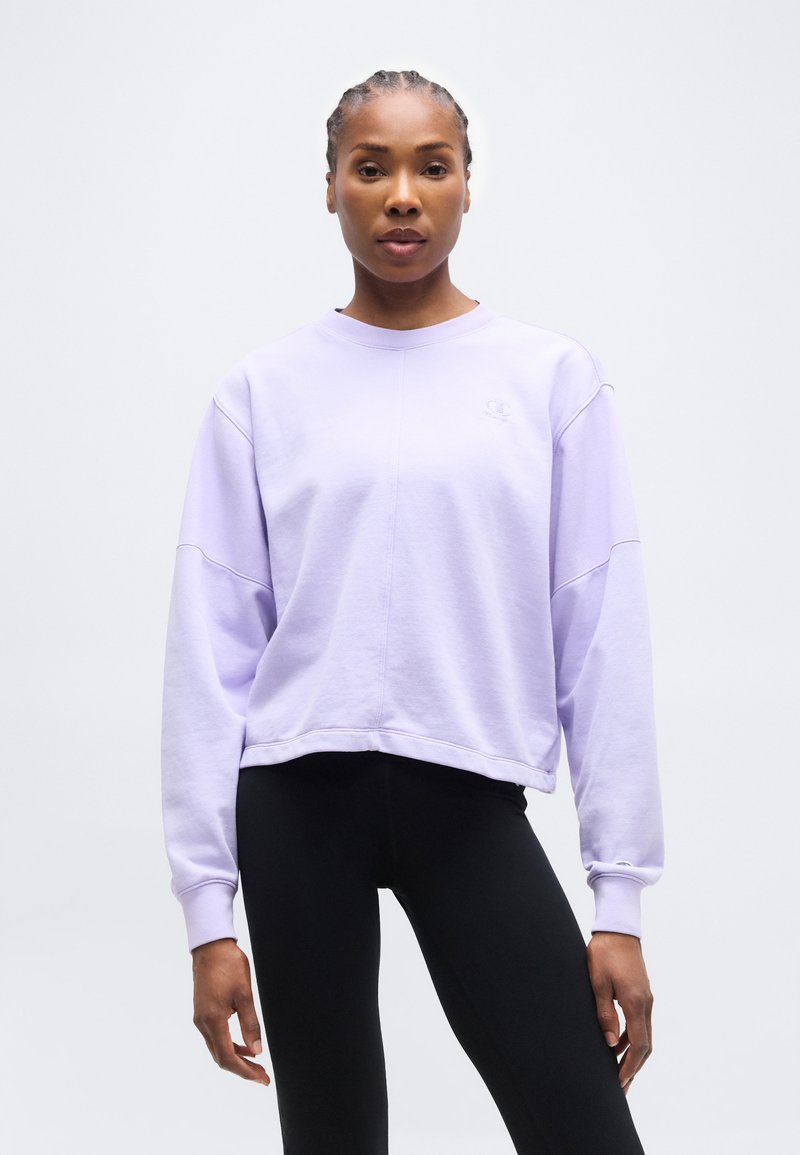 Γυναίκα με πλεξούδες φοράει ένα φαρδύ φούτερ ανοιχτό μωβ και μαύρο κολάν, στέκεται μπροστά σε ένα απλό γκρι φόντο.