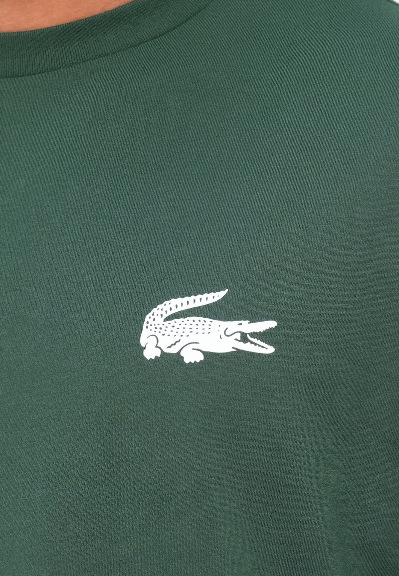 Grünes Baumwoll-T-Shirt mit einem weißen Krokodil-Logo auf der linken Brust. Weicher Stoff mit Rundhalsausschnitt.