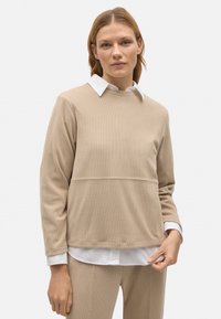 Top beige de manga larga con textura acanalada, con un escote redondo y una camisa blanca debajo, combinado con pantalones beige a juego.