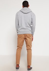 Uomo con felpa grigia, pantaloni beige e sneakers grigie si trova di fronte a un muro bianco semplice con le mani rilassate ai lati.