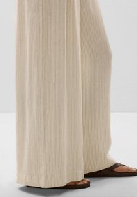 Light beige wide-leg pants with vertical stripes, made of textured fabric. Paired with brown flat sandals, providing a casual look.