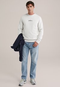 White sweatshirt with black printed text, light blue jeans, and gray sneakers. A navy jacket is draped over the arm. Neutral background.