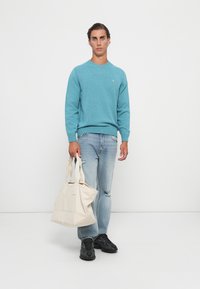 Mann in einem türkisen Pullover und hellblauen Jeans, der eine große weiße Tragetasche hält, steht auf einem weißen Boden vor einer schlichten weißen Wand.