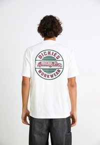 Hvid bomulds T-shirt med et cirkulært grafisk design på ryggen, med "DICKIES" og en illustration af en lastbil, accentueret i grøn og maroon.