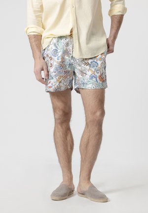 Hombre con camisa amarilla de manga larga, pantalones cortos coloridos con estampado floral y zapatos grises sin cordones, de pie con las manos relajadas.