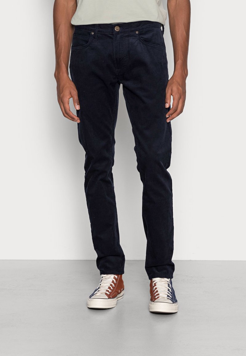 Jean en velours côtelé bleu marine avec une coupe slim, dotés de poches avant et d'une fermeture à bouton en métal. Portés avec des baskets marron et bleues.